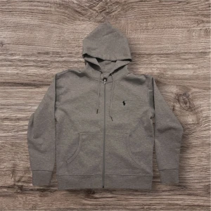 Grå hoodie från Polo Ralph Lauren - Snygg grå hoodie från Polo Ralph Lauren med dragkedja och klassisk logga broderad på bröstet. Tröjan har huva med snörning, två fickor fram och ribbade muddar. Perfekt för chill dagar och enkel att matcha med jeans eller joggers.