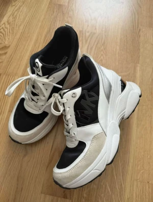 Chunky sneakers från Michael Kors - Säljer ett par chunky sneakers från Michael Kors i svart, vitt och beige med silvriga detaljer. Skorna har höga sulor, snörning och stora MK-loggor på sidorna. Materialmix av skinn, mocka och syntet ger en lyxig och sportig vibe. Perfekta för dig som gillar statement-sneakers.