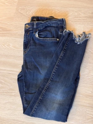 Mörkblå jeans från River Island - Snygga mörkblå jeans från River Island med slitna och fransade benslut. Klassisk femficksmodell och normal passform. Perfekt för dig som gillar en lite ruffare stil med coola detaljer.