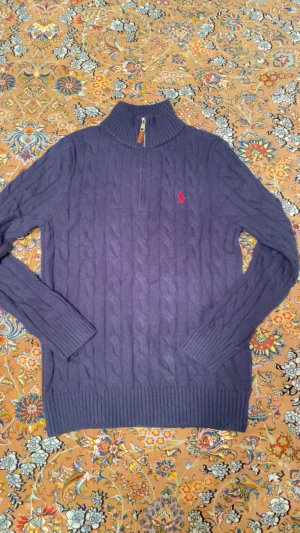 Polo Ralph Lauren Tröja - Snygg mörkblå polotröja från Polo Ralph Lauren med klassisk kabelstickad design och röd broderad logga på bröstet. Tröjan har half zip med läderdetalj och ribbade muddar vid ärmslut och nederkant. Perfekt för dig som gillar stilren och tidlös look. Står M men sitter som S