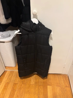 Svart lång dunväst med huva - Svart quiltad dunväst med huva och dragkedja framtill. Västen har två fickor på sidorna och en stilren, enkel design som passar perfekt över en hoodie eller tröja. Lång modell som ger extra värme och streetkänsla.