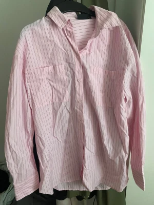 Rosa randig skjorta från Pieces XS - Säljer en rosa skjorta från Pieces i storlek XS. Skjortan har tunna svarta ränder, klassisk krage, knäppning framtill och två bröstfickor. Långärmad och tillverkad i mjuk bomull. Perfekt för en chill och stilren look.