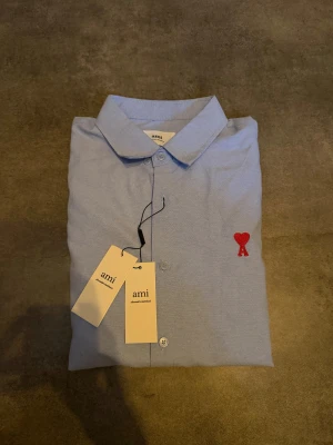 Ljusblå skjorta från Ami Paris - Snygg ljusblå skjorta från Ami Paris med klassisk krage och knäppning framtill. Skjortan har ett rött broderat hjärta med bokstaven A på bröstet. Tillverkad i bomull och har långa ärmar. Perfekt för dig som gillar stilrena detaljer. Står XL men passar som L