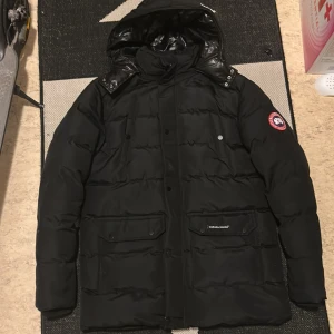 Svart dunjacka från Canada Goose L - Säljer en svart dunjacka från Canada Goose i storlek L. Jackan har huva, flera fickor med lock och tryckknappar samt klassisk Canada Goose-logga på ärmen. Perfekt för kalla vinterdagar och riktigt snygg streetstil.