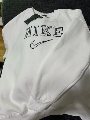 Vit sweatshirt från Nike XL - Vit sweatshirt från Nike i storlek XL med broderad logga och swoosh på bröstet. Tröjan har rund hals, långa ärmar och ribbade muddar vid ärmslut och nederkant. Perfekt för en chill och sportig look. Aldrig använd, säljer pga stor i storlek 