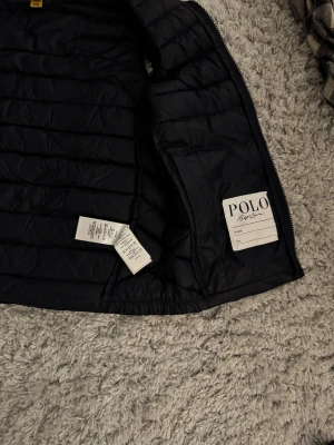 Svart dunväst från Polo Ralph Lauren - Svart quiltad dunväst från Polo Ralph Lauren med ståkrage och dragkedja framtill. Västen har två fickor med dragkedja och den klassiska röda Polo-loggan broderad på bröstet. Perfekt för lager-på-lager och snygg till streetwear.
