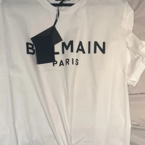 Vit t-shirt från Balmain Paris - Snygg vit t-shirt från Balmain Paris med svart tryck framtill. Klassisk rund hals och korta ärmar. Perfekt för dig som gillar exklusiva märken och stilren design. Materialet känns mjukt och skönt mot huden.