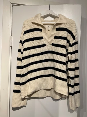 Randig stickad tröja från H&M - Säljer en krämvit och svart stickad tröja från H&M med breda ränder. Tröjan har en djup v-ringning, stor krage och långa ärmar. Den är ribbad vid muddar och nederkant, vilket ger en chill och snygg look. Perfekt för lager-på-lager under hösten. Är i ny skick