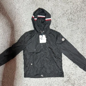 Svart  windbreaker från Moncler med huva - Svart vindjacka från Moncler med huva och dragkedja framtill. Jackan har Moncler-logga på ärmen och snygga röda och vita ränder på huvan. Tillverkad i lätt polyester, perfekt för blåsiga dagar. Två fickor med tryckknappar och justerbar dragsko vid huvan.