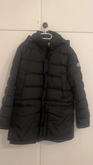 Svart Moncler dunjacka med päls - Säljer en svart dunjacka från Moncler med klassisk logga på ärmen. Jackan har huva med brun pälsdetalj, stora fickor framtill och dragkedja. Perfekt för kalla dagar och riktigt snygg streetstil.