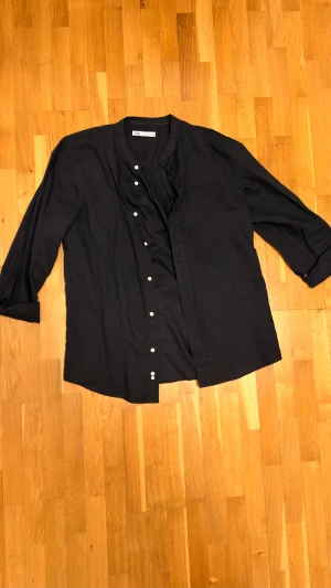 Svart skjorta från Zara - Svart henley-skjorta från Zara i avslappnad passform. Skjortan har ståkrage, vita knappar framtill och långa ärmar. Tillverkad i ett mjukt bomullstyg som känns skönt mot huden. Perfekt för en stilren och enkel look.