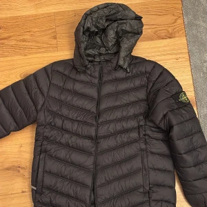 Svart dunjacka från Stone Island - Svart dunjacka från Stone Island med avtagbar huva och klassisk patch på vänster ärm. GÅR ATT SÄTTA PÅ ELLER TA AV LUVAN. 