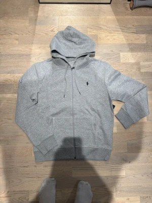Grå hoodie från Polo Ralph Lauren - Snygg grå hoodie från Polo Ralph Lauren med klassisk logga broderad på bröstet. Storlek M. Han på bild är 185cm, 74kg. Stilren och enkel att matcha med jeans eller joggers. Sista bilden är hemsidan man kommer till när man scannar QR koden.