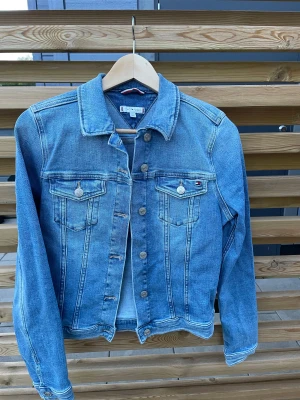 Blå jeansjacka Tommy Hilfiger 36 - Snygg klassisk blå jeansjacka från Tommy Hilfiger i storlek 36. Jackan har raka linjer, två bröstfickor med knappar och ikonisk logga på ena fickan. Tillverkad i jeansmaterial med silverfärgade knappar och krage. Perfekt för dig som gillar en tidlös streetstil.