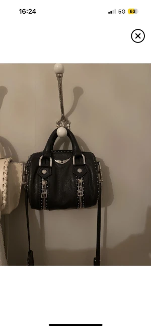 Zadig väska  - Cool svart handväska i skinn med silverfärgade metalldetaljer och dragkedja upptill. Väskan har två handtag och en liten silverlogga framtill. Perfekt storlek för det viktigaste och ger en edgy vibe till din outfit. Zadig väska 