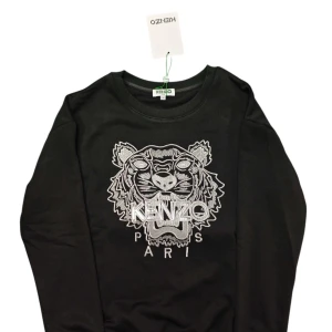 Svart sweatshirt med tiger från Kenzo - Svart sweatshirt från Kenzo i storlek M med ikonisk broderad tiger och Kenzo Paris-logga i silver på bröstet. Tröjan har rund hals, långa ärmar och är tillverkad i mjuk bomull. Perfekt för dig som vill ha en statement piece med streetstyle-vibe.