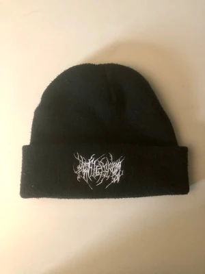 Svart stickad mössa med broderi - Svart mössa i stickad material med uppvikt kant och ett coolt vitt broderat mönster framtill. Perfekt för dig som gillar streetstyle och vill ha något som sticker ut. Broderiet har en edgy, grafisk stil som ger mössan en unik vibe.