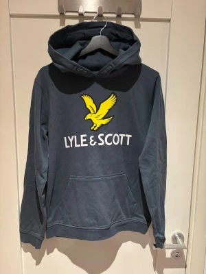 Blå/grön hoodie från Lyle & Scott  - Snygg hoodie i färgen Orion Blue från Lyle & Scott Junior med stor gul fågel och vit logga på bröstet. Tröjan är inte använd. Materialet är mjuk bomull blandat med polyester, perfekt för chill dagar.