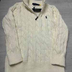 Vit kabelstickad tröja Polo Ralph Lauren - Vit kabelstickad tröja från Polo Ralph Lauren med halv dragkedja och broderad logga på bröstet. Tröjan har ribbade muddar och hög krage, perfekt för kyliga dagar. Klassisk och stilren design som funkar till allt. Påse följer med.