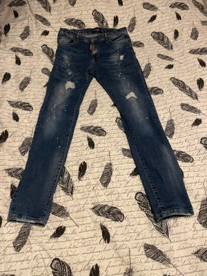 Blå Dsquared2 jeans med slitningar - Säljer ett par blå jeans från Dsquared2 i storlek 158. Jeansen har coola slitningar och färgstänk på benen, klassisk femficksmodell och Dsquared2-logga på röd etikett vid gylfen. Materialet är 98% bomull och 2% elastan för en skön passform.