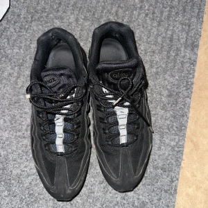 Svarta Nike Air Max 95 - Säljer ett par svarta Nike Air Max sneakers med mesh och syntetdetaljer. Skorna har snörning och en platt sula med tydlig dämpning. Perfekta för dig som gillar sportig stil och vill ha bekväma skor till vardagen.