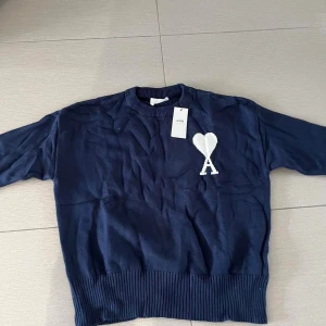 Mörkblå stickad tröja från Ami - Snygg mörkblå stickad tröja från Ami med vit hjärtlogga och bokstaven A på bröstet. Tröjan har rund hals, långa ärmar och bred ribbad kant vid midja och ärmslut. Perfekt för dig som gillar stilrena detaljer och vill ha något som sticker ut.