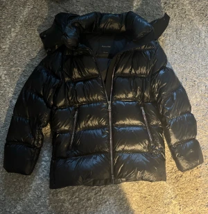 Svart pufferjacka från Massimo Dutti använd endast 5 gånger - Säljer en svart pufferjacka från Massimo Dutti med glansig finish och hög krage. Jackan har två snedställda fickor med dragkedja, avtagbar huva och breda kanaler. Perfekt för kalla dagar. Har endast använt jackan 5 gånger så jackan är i nyskick. Passar bra på mig som är 165 cm. Skulle säga att xs även passar 170 cm.