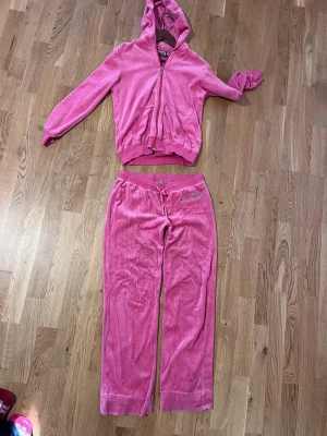 Vintage Juicy Couture rosa set - Rosa set från Juicy Couture med strassdetaljer på höften och snörning i midjan. Byxorna har en loose passform och lågmidjade och är tillverkade i mjuk velour som känns skön mot huden. Perfekt för chill dagar och har en ikonisk 00-tals vibe. Koftan är jätte skön o fin passform. Står storlek XL men passar S!!