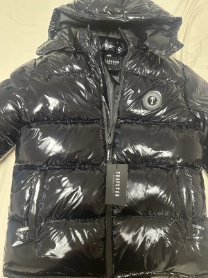 Svart pufferjacka från Trapstar - Snygg svart pufferjacka från Trapstar med glansig finish och stor huva. Jackan har dragkedja framtill, två fickor och Trapstar-logga på bröstet samt broderad logga på ryggen. Perfekt för kalla dagar och riktigt streetwear-vibe.