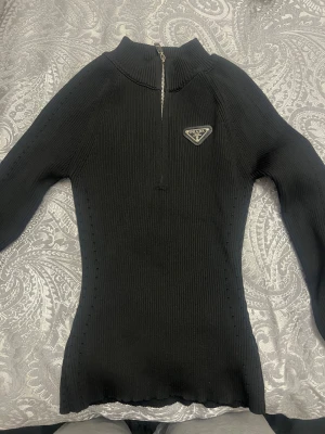 Svart polotröja Prada - Svart polotröja från Prada med ribbad struktur och halv dragkedja framtill. Tröjan har lång ärm och en snygg logotyp på bröstet. Passformen är slim och materialet känns mjukt och stretchigt, perfekt för att lyfta din stil.