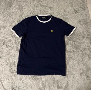 Marinblå t-shirt från Lyle & Scott XL - Snygg marinblå t-shirt från Lyle & Scott i storlek XL. T-shirten har vita detaljer vid krage och ärmslut samt den klassiska gula fågeln broderad på bröstet. Tillverkad i mjuk bomull och har en normal passform med korta ärmar.