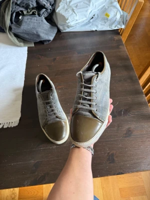 Lanvin grå sneakers med brunt skinn - Lanvin sneakers i grå mocka med bruna skinn-detaljer på tån och runt snörningen. Klassisk rund tå och platt vit sula med logotyp undertill. Snygg kontrast mellan material och färger, perfekt för dig som gillar stilrena och exklusiva sneakers.