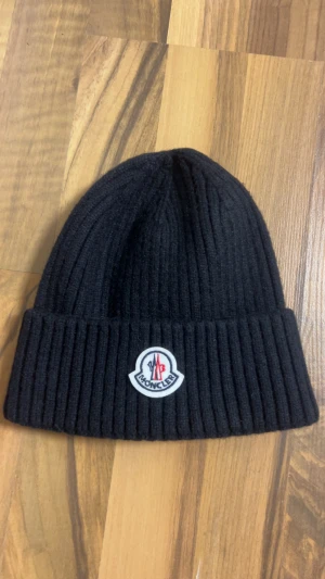Svart ribbad Moncler mössa - Svart ribbstickad mössa från Moncler med klassisk uppvikt kant och broderad logotyp framtill. Snygg och enkel design som ger en clean look, perfekt för dig som gillar exklusiva märken och vill hålla dig varm med stil.