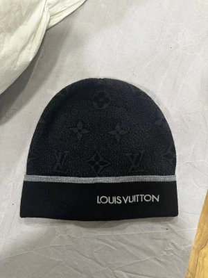 Svart mössa i ull med logotyp  - Snygg svart mössa med klassiskt monogrammönster och silverfärgad rand. Broderad logga framtill. Tillverkad i 100% ull, perfekt för kalla dagar när du vill ha både stil och värme.