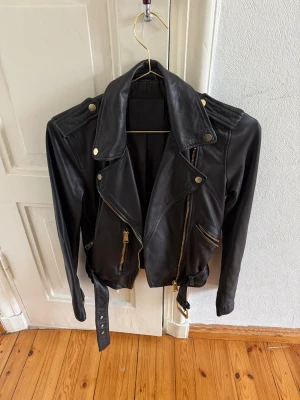Svart skinnjacka AllSaints biker - Snygg svart skinnjacka från AllSaints i klassisk bikerstil med guldiga dragkedjor, axelklaffar och bälte i midjan. Jackan har flera fickor med dragkedja och coola detaljer som ger en edgy vibe. Perfekt för dig som vill ha en tidlös och stilren look.