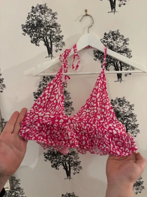 Rosa leopardmönstrad topp från Express - Snygg topp från Express i rosa och vitt leopardmönster. Modellen har volangkant nedtill och knyt i nacken för justerbar passform. Materialet är lätt och syntetiskt, perfekt för strandhäng. Storlek XS.