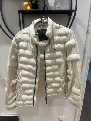 Vit dunjacka från Morris, M - Säljer en fräsch vit dunjacka från Morris i storlek medium. Jackan har quiltad design, svart dragkedja och står ut med sin stilrena look. Perfekt för dig som vill ha en lätt och snygg jacka med hög krage och diskret logga på bröstet.