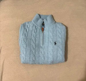 Ljusblå stickad tröja Polo Ralph Lauren - Har använts typ 3-4 gånger, en Snygg ljusblå stickad tröja från Polo Ralph Lauren med klassiskt kabelmönster och half zip-dragkedja med läderdetalj. Tröjan har ribbad krage, muddar och nederkant samt långärmad passform. Perfekt för dig som gillar stilrena och tidlösa plagg.