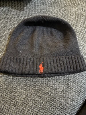 Svart stickad mössa från Polo Ralph Lauren - Svart ribbstickad mössa från Polo Ralph Lauren med klassisk röd broderad logga framtill. Mjuk och skön passform, perfekt för kyliga dagar. Enkel design som funkar till de flesta stilar.