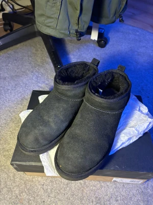 Svarta UGG boots low  - Säljer ett par svarta UGG boots med rund tå och platt sula. Utsidan är i mjuk mocka och insidan är fodrad med fluffigt material för extra värme. Skorna har en låg siluett och UGG-logga baktill. Perfekta för kalla dagar och har en klassisk look.