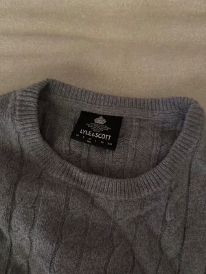 Grå kabelstickad tröja Lyle & Scott - Snygg grå kabelstickad tröja från Lyle & Scott med rund hals och lång ärm. Tröjan har ett broderat gult logomärke på bröstet och ribbade muddar vid hals, ärmslut och nederkant. Perfekt för dig som gillar klassisk stil med en modern touch.