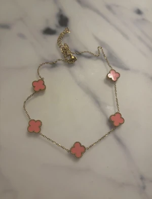 Guldkedja med rosa fyrklöver - Snyggt halsband med tunn guldkedja och fem rosa fyrklöver-formade hängen. Hängena har en fin guldfärgad kant som ger extra detalj. Perfekt för dig som gillar färg och unika former.