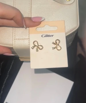 Guldiga rosettörhängen från Glitter - Supersöta örhängen i form av små rosetter i guldig färg från Glitter. Perfekt för dig som vill ha en gullig och feminin touch på din look. Rosettformen ger en lekfull vibe och de är enkla att matcha med andra smycken.