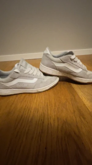 Vita Vans sneakers  - Säljer ett par stilrena Vans sneakers i ljusgrått och vitt. Skorna har klassisk snörning, rund tå och platt sula med gummimönster undertill. Ovandelen är i mjuk mocka med den ikoniska Vans-sidan i vitt. Perfekta för dig som gillar streetstyle och skate.