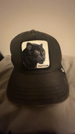 Svart keps med pantermärke - Cool svart keps från Goorin Bros med broderad patch framtill som visar en panter och texten 'Panther'. Kepsen har böjd skärm och justerbar baksida. Perfekt för dig som vill sticka ut med djurmotiv.