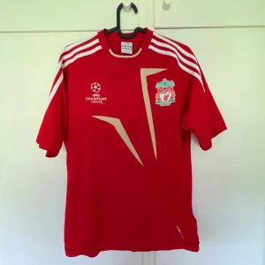 Liverpool Champions League tröja Adidas - Röd Liverpool fotbollströja från Adidas med Champions League-logga och klubbmärke på bröstet. Tröjan har vita detaljer, guldiga grafiska inslag och klassiska Adidas-ränder på axlarna. Tillverkad i lätt och ventilerande material, perfekt för match eller träning.