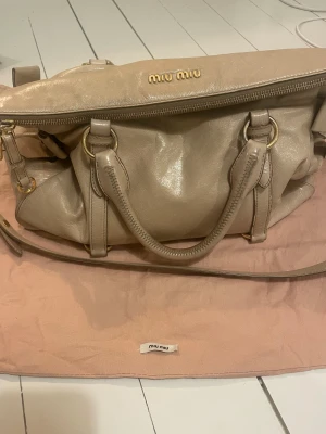Beige handväska från Miu Miu i skinn - Snygg beige handväska från Miu Miu i mjukt skinn med guldfärgade metalldetaljer och dragkedja. Väskan har både handtag och axelrem samt en dekorativ rosett på sidan. Perfekt för dig som vill ha en lyxig och stilren accessoar. Kan gå ner i pris vid snabb affär 🫶🫶