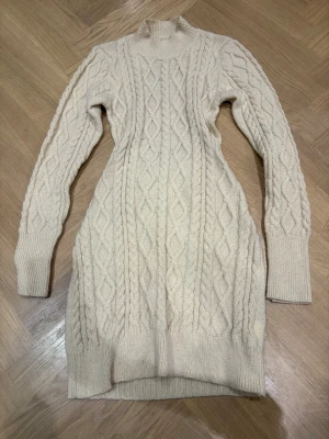 Stickad beige klänning med öppen rygg - Supermysig stickad klänning i beige med flätstickat mönster och hög krage. Klänningen har lång ärm och en cool öppen ryggdetalj som gör den extra unik. Passformen är normal och materialet känns mjukt och varmt, perfekt för kyliga dagar.