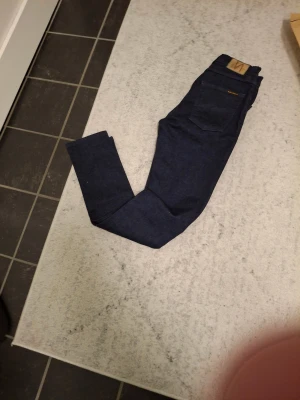 Mörkblå nudie jeansbyxor W26 L32 - Snygga mörkblå jeansbyxor i klassisk rak modell. Jeansen har fem fickor, läderpatch bak i midjan och är tillverkade i slitstarkt denim. Perfekta för dig som gillar stilrena och tidlösa jeans med en clean look.