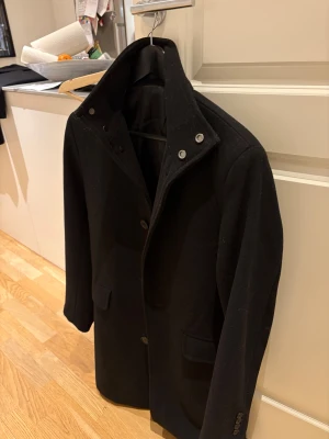 Svart rock (HM) hög krage, knappar - Snygg svart rock från HM med stilren design och hög krage. Jackan har knappar framtill och två fickor med lock. Passar perfekt för dig som gillar en klassisk look med lite edge. Materialet är mjukt och känns varmt, perfekt för kyliga dagar.